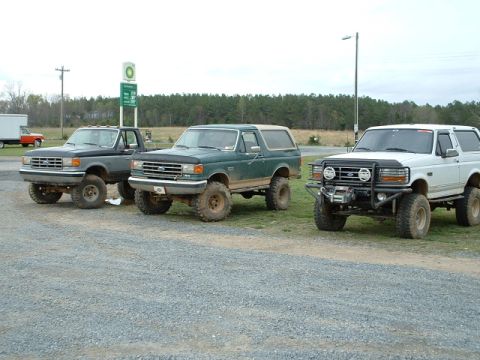 1987 Ford F-150 - Three lifted Ford Broncos from the late 1980s are parked side-by-side in a gravel lot.