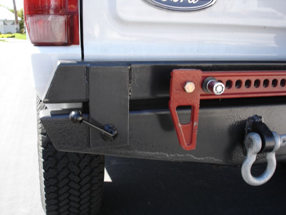 Custom welded steel rear bumper on a light blue Ford truck with red Hi-Lift jack mount and D-ring shackle