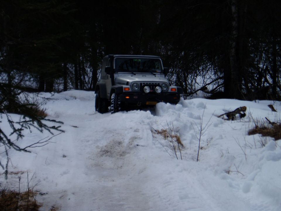 Silver Jeep Wrangler driving through deep snow, front view. Headlights on, aftermarket bumper