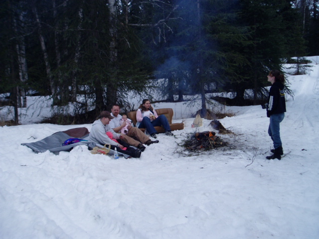 people sitting in the snow around a campfire, with trees in the background