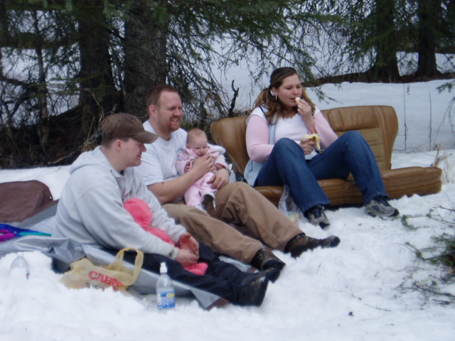 People sitting in the snow around a couch, with a grocery bag and water bottle visible