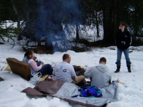 2000 Ford E350 - A group of people enjoy a campfire in a snow-covered wooded area, with one person standing in the background.