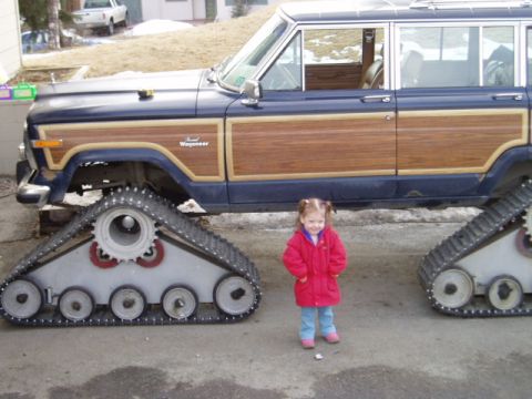 2000 Ford E350 - A young child stands beside a modified Jeep Grand Wagoneer, which has been equipped with tank treads for off-road capability.