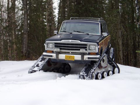 2000 Ford E350 - A Jeep Grand Wagoneer modified with snow tracks navigates a snow-covered terrain surrounded by trees.