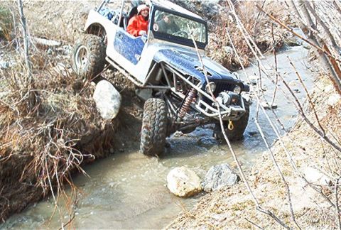 1986 Ford Bronco - Off-road Jeep Wrangler tackles a challenging stream crossing with large tires and suspension modifications.