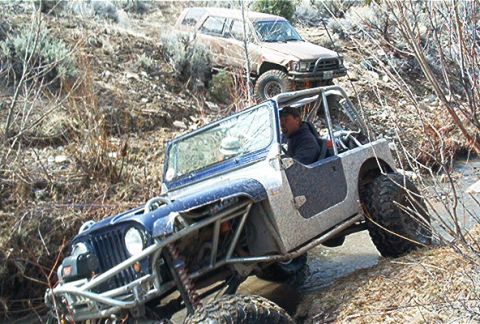 1986 Ford Bronco - Two off-road vehicles traverse a challenging, watery terrain with dense brush.