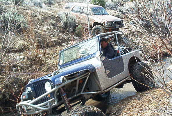Modified Jeep Wrangler with roll cage navigating a rocky stream, a tan SUV behind it