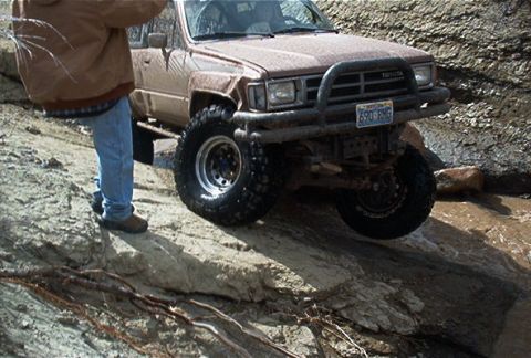 1986 Ford Bronco - Toyota 4Runner tackling a challenging off-road course with large tires and a modified bumper.