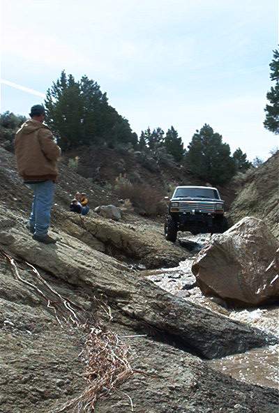 1986 Ford Bronco - A person stands on a rocky incline watching a 4x4 vehicle navigate a shallow, rocky stream.