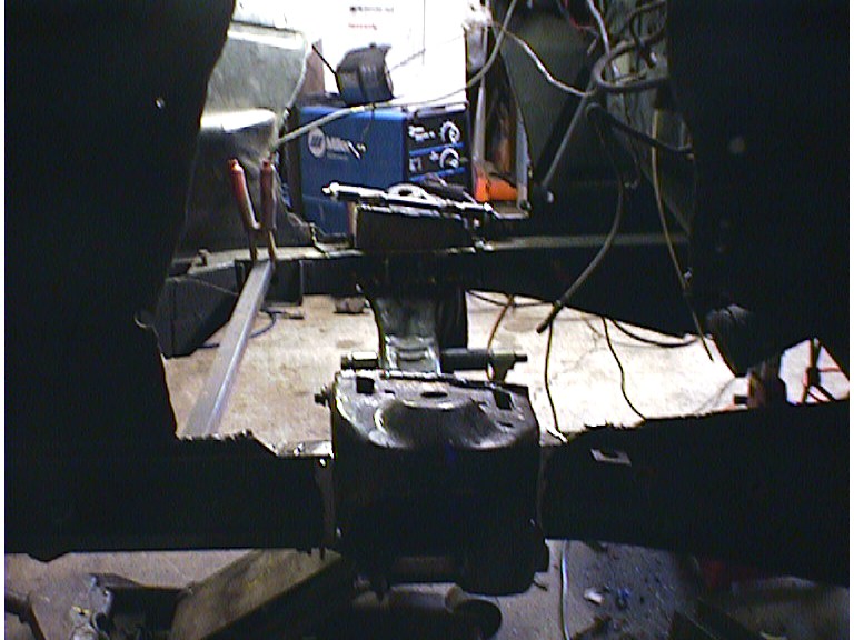 Bare vehicle frame in a dim shop with a driveline component between the rails and a Miller welder behind