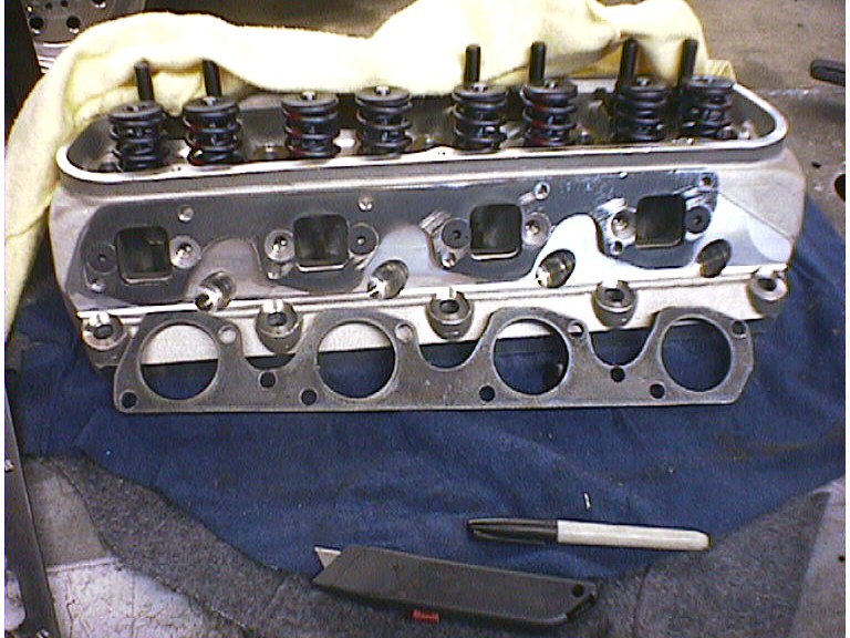 Aluminum V8 cylinder head with valve springs installed and an exhaust flange gasket leaning against its ports