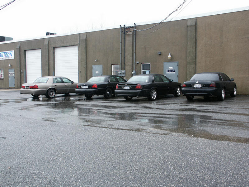 Four sedans, three black and one tan, parked in a row outside a building. The rear of the vehicles is visible