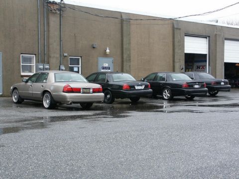 2001 Mercury Grand Marquis - Four sedans, including a tan 2001 Mercury Grand Marquis, are parked outside a commercial building on a wet day.