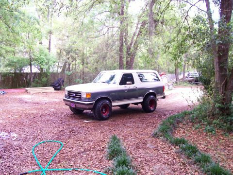 1990 Ford Bronco - A two-tone gray and white Ford Bronco with red wheels sits parked on a gravel driveway surrounded by trees.