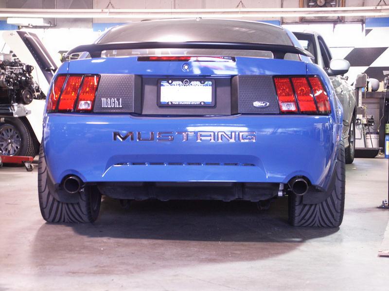 Rear view of a blue Ford Mustang Mach 1 with wide tires, showing "MACH 1" and "MUSTANG" badging