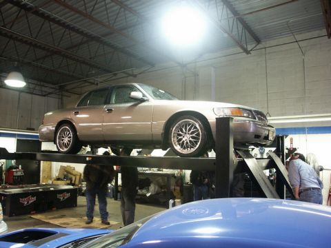 2001 Mercury Grand Marquis - A tan Mercury Grand Marquis is raised on a car lift inside a workshop, with another vehicle partially visible in the foreground.