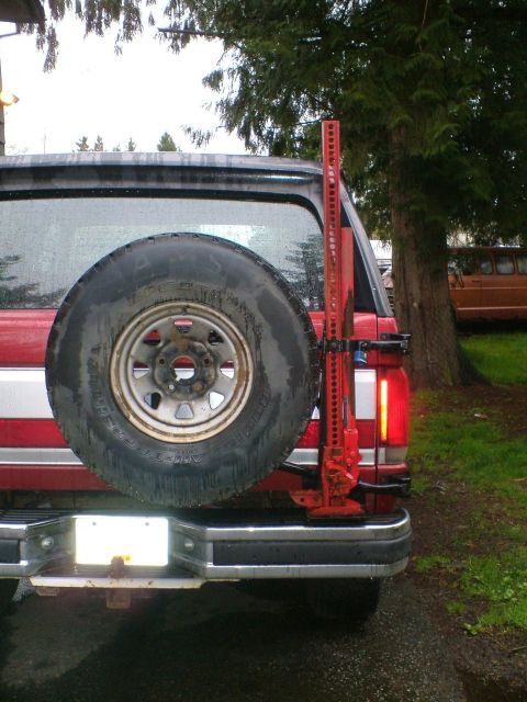 1989 Ford Bronco - Straight-on rear view of a rain-soaked red and white Ford Bronco with a swing-out tire carrier holding a BFGoodrich All-Terrain T/A spare o…