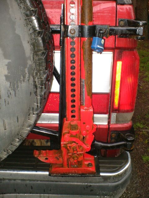 1989 Ford Bronco - A red Hi-Lift-style jack is mounted vertically on a rear tire carrier and secured with a blue Master padlock on a dark red Ford Bronco. The…