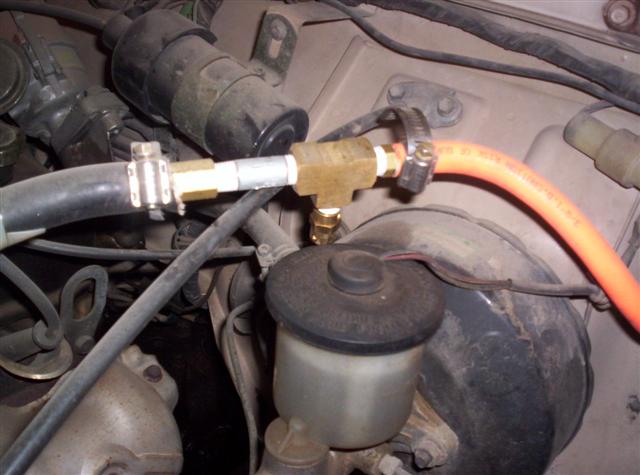Engine bay showing a brass T-fitting connected to hoses and a brake fluid reservoir