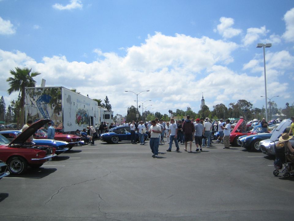 Outdoor car show with many classic cars parked and people milling around. A large white trailer with graphics is on the left