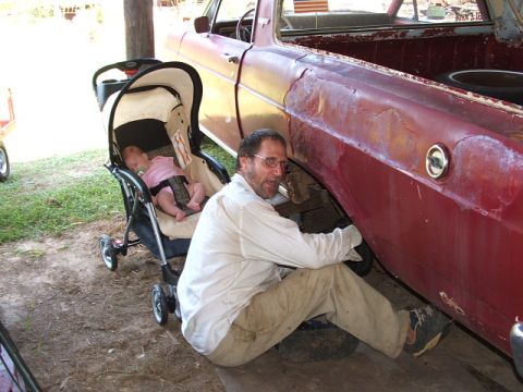 1966 Ford Ranchero - A man works on the side of a red vehicle while a baby sleeps in a stroller nearby.