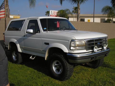1966 Ford Bronco - Three-quarter front view of a white 1990s full-size Ford Bronco parked on grass, lifted on mud-terrain tires with polished five-spoke alloy…