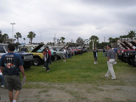1966 Ford Bronco - Outdoor car show on a grass field with two long rows of early Ford Broncos parked hoods-up, including yellow, green, red and dark-colored t…