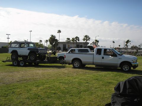 1966 Ford Bronco - A white and dark blue 1966 Ford Bronco is transported on a trailer by a white pickup truck under a partly cloudy sky.