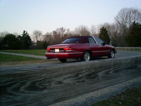 1995 Ford Crown Victoria - A red sedan with a vinyl top is seen from the rear on a road with trees in the background.