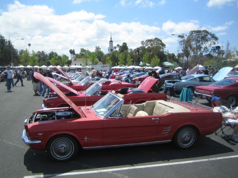 1968 Ford Mustang - Multiple classic Ford Mustangs are displayed at an outdoor car show under a partly cloudy sky.