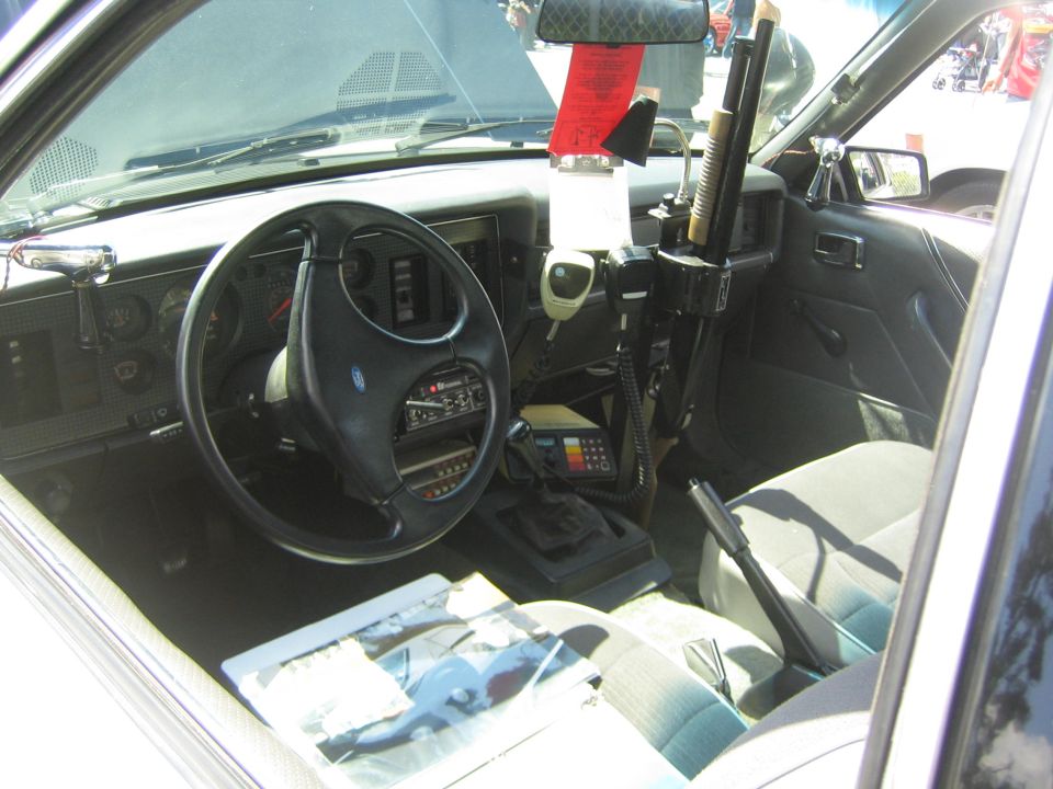 Car interior shows steering wheel, radio, shotgun mounted on dash, and a red ticket on the mirror