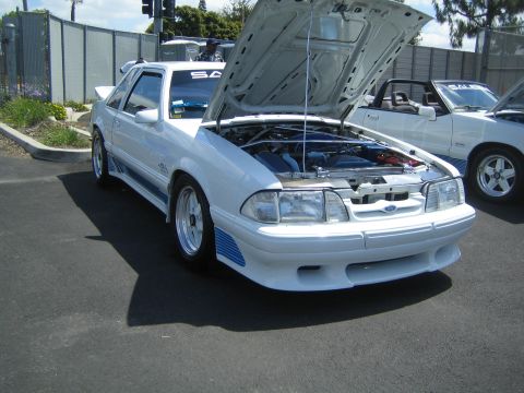 1968 Ford Mustang - A white 1987 Ford Mustang with blue racing stripes is parked outside with its hood open, revealing the engine.