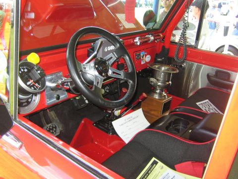 1968 Ford Mustang - The interior of a red vehicle features racing seats, a custom steering wheel, and a trophy, with a document on the floor.