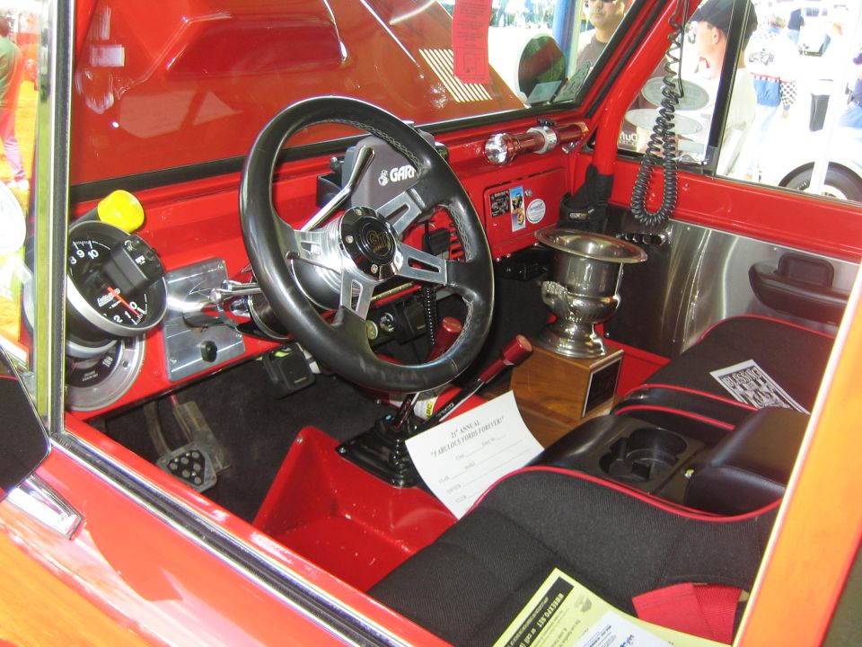 Interior of a red vehicle with a custom steering wheel, racing seats, and a trophy on the center console. A document is visible on the floor