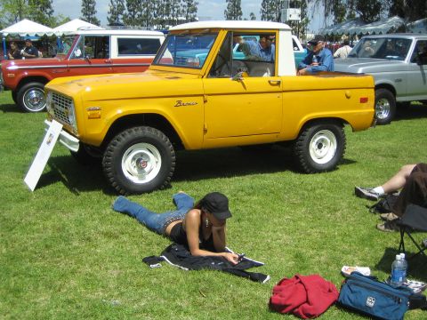 1968 Ford Mustang - A yellow 1975 Ford Bronco with a white top is displayed at an outdoor car show, with a woman relaxing on the grass in the foreground.