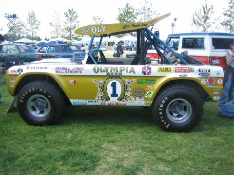 1968 Ford Mustang - A heavily modified off-road vehicle with "Big Oly" branding is displayed on a grassy field with other vehicles in the background.