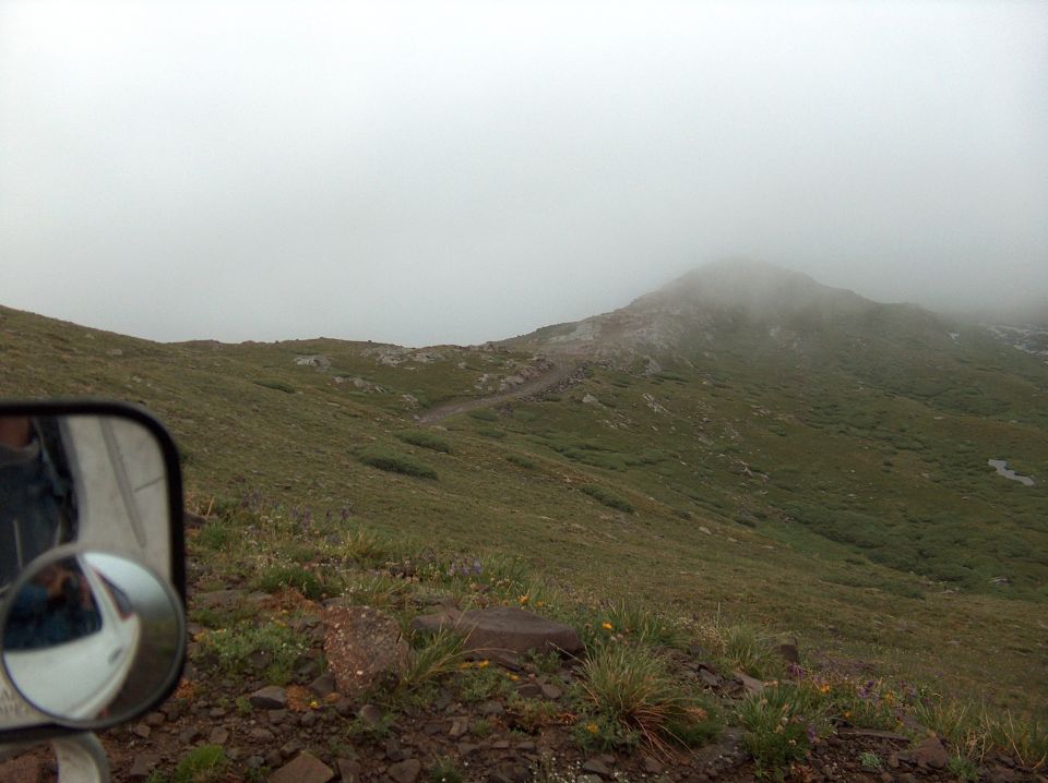 Side mirror reflecting a white vehicle, with a foggy alpine trail and mountainside ahead