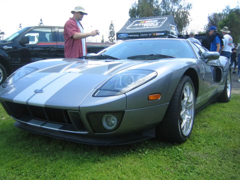 1968 Ford Mustang - A gray Ford GT with silver racing stripes is parked on grass, with a man taking a photo and a NASCAR tent in the background.