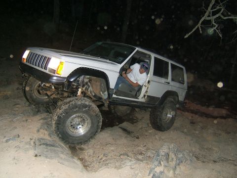 1993 Ford Bronco - A man drives a modified Jeep Cherokee XJ over rough terrain in the dark.