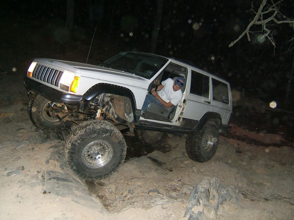 White Jeep Cherokee XJ with lifted suspension and large tires navigating a rocky trail at night