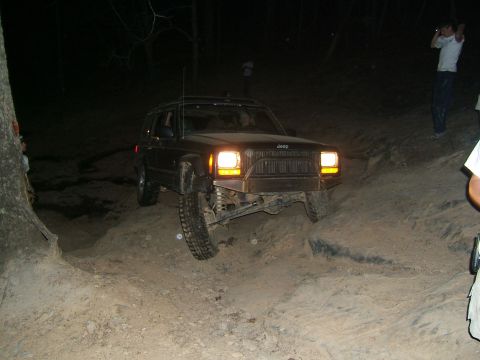 1993 Ford Bronco - A dark Jeep Cherokee XJ navigates a challenging off-road trail in the dark, with its headlights illuminating the path ahead.