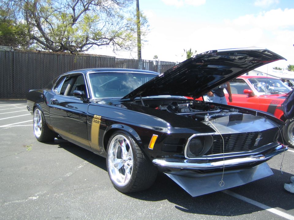 Black 1970 Ford Mustang Boss 302 with hood open, gold stripe on side. Red car behind it