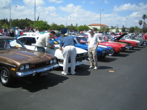 1968 Ford Mustang - A line of classic cars are parked at an outdoor event, with people gathered around them.