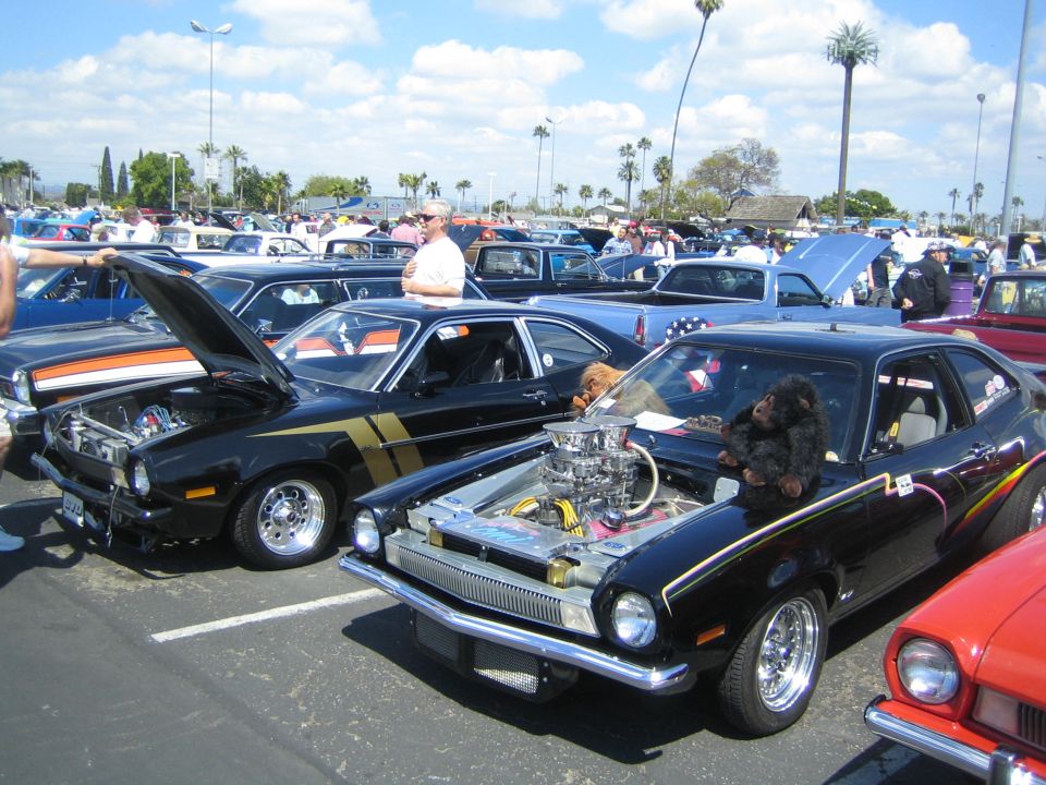 Black modified Ford Pinto with gold stripes, engine exposed, and a stuffed monkey on the hood. Another Pinto is next to it
