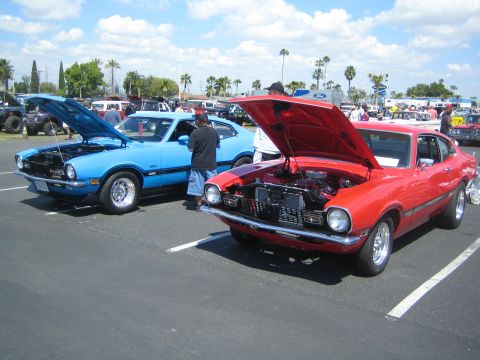 1968 Ford Mustang - Two classic Ford Mavericks, one red and one blue, are displayed with their hoods open at an outdoor car show.