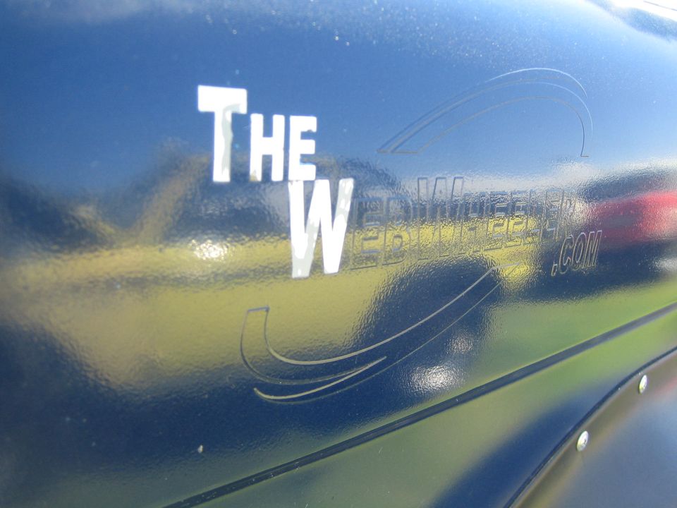 Side of a dark blue vehicle with white text "THE W" and a graphic. Reflected text reads "THE WEBCOMPILER.COM"