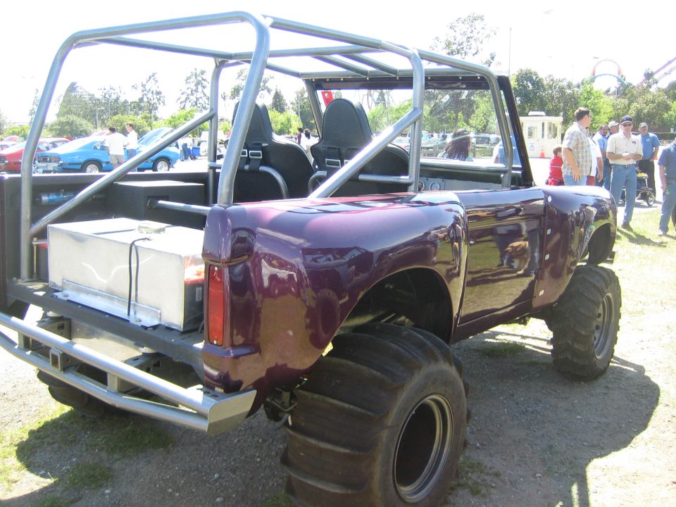Purple custom off-road vehicle with roll cage, large tires, and a fuel tank in the bed. Other cars and people are visible in the background