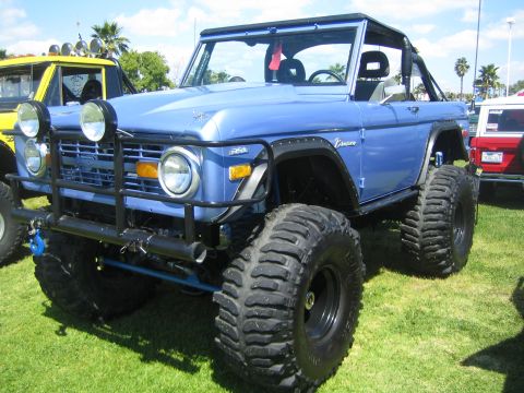 1968 Ford Mustang - A lifted blue Ford Bronco with oversized tires sits on a grassy field, parked next to other vehicles.
