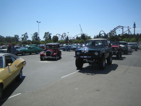 1968 Ford Mustang - A line of classic vehicles, including a black Ford Bronco, are parked in a lot with a roller coaster in the background.