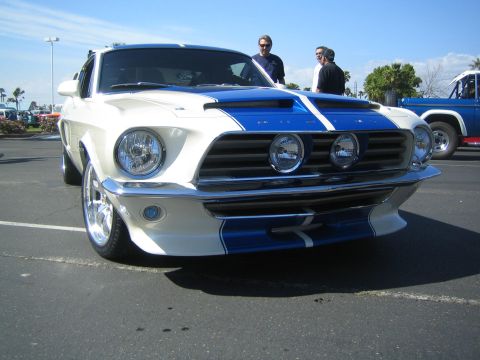 1968 Ford Mustang - A white 1968 Ford Mustang with blue racing stripes is parked at an outdoor event, with other vehicles and people in the background.